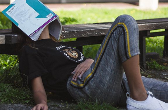 Student leaning against bench with book resting on face exhausted of studying